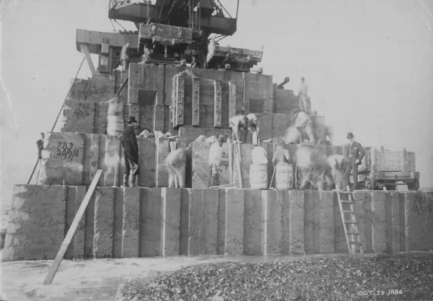 Workers on a stepped pile of numbered concrete blocks at the start of Roker Pier, with a crane behind and barrels at their feet.