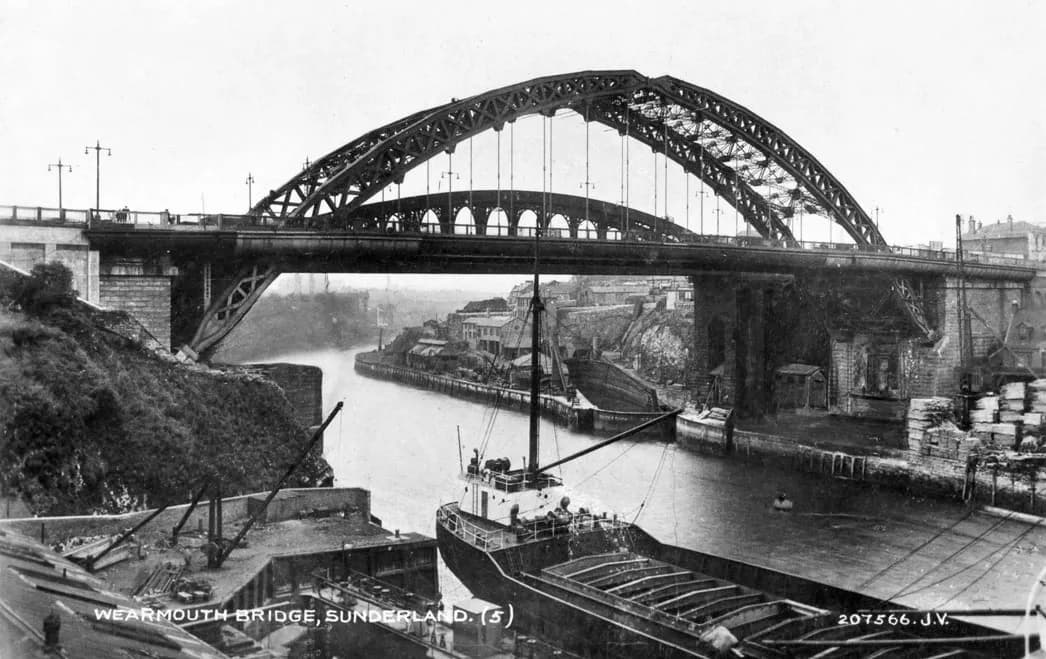 The steel arch of Wearmouth Bridge over the River Wear, with a collier moored in the foreground and quays and houses beyond.