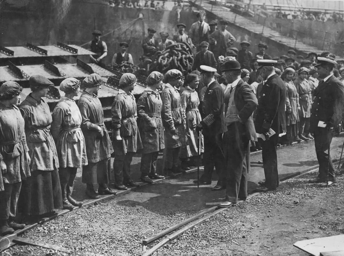 A line of women shipyard workers in caps and overalls meet King George V in naval uniform, with men watching from the slipway behind.